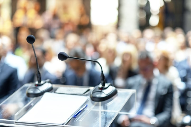 Two microphones, paper, and a pen in focus on a podium with a blurred background of audience members