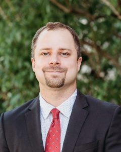 Dr. Ashton Verdery, a man with light brown hair who is wearing a suit with a red tie.